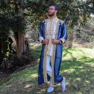 May include: A navy blue robe with gold embroidery and trim, worn over a white outfit. The robe features intricate gold detailing along the edges and sleeves. The outfit is completed with light blue slippers. The background is a natural outdoor setting.