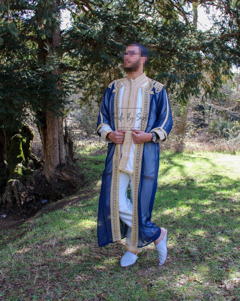 May include: A navy blue robe with gold embroidery and trim, worn over a white outfit. The robe features intricate gold detailing along the edges and sleeves. The outfit is completed with light blue slippers. The background is a natural outdoor setting.