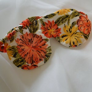 Könnte beinhalten: Eine florale Schlafmaske mit orangefarbenen, gelben und grünen Blumen auf weißem Hintergrund.