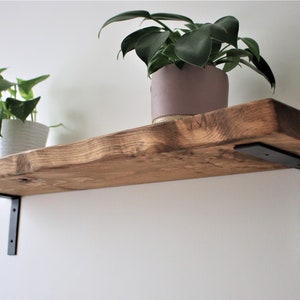May include: A wooden shelf with a natural finish and black metal brackets. Two potted plants are on the shelf. The shelf is mounted to a white wall.