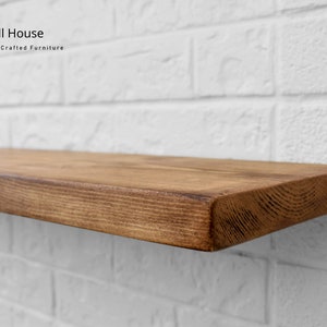 Op de afbeelding: Een rustiek houten plank met een natuurlijke bruine afwerking. De plank is gemonteerd op een witte bakstenen muur. Chill House Handgemaakte meubels.