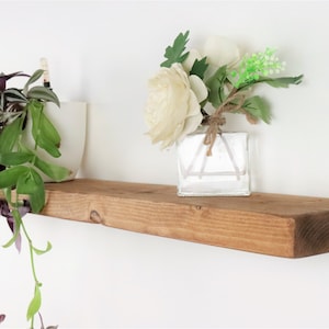 May include: A rustic wooden shelf with a natural brown finish. The shelf is decorated with a white ceramic pot with a trailing plant and a clear glass vase with white flowers.