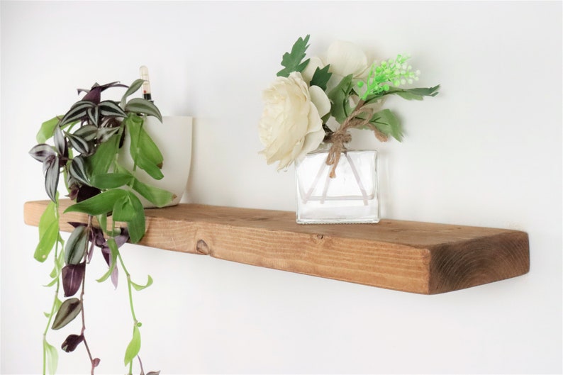 May include: A rustic wooden shelf with a natural brown finish. The shelf is decorated with a white ceramic pot with a trailing plant and a clear glass vase with white flowers.