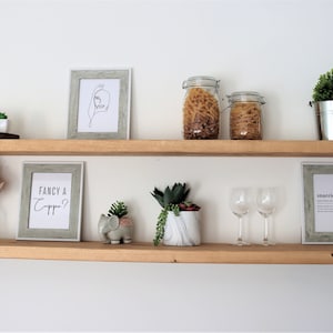 May include: Two wooden shelves with various items on them, including a framed print that says "Fancy a Capp?", a small elephant figurine, two glass jars filled with pasta, two wine glasses, and several small potted plants.