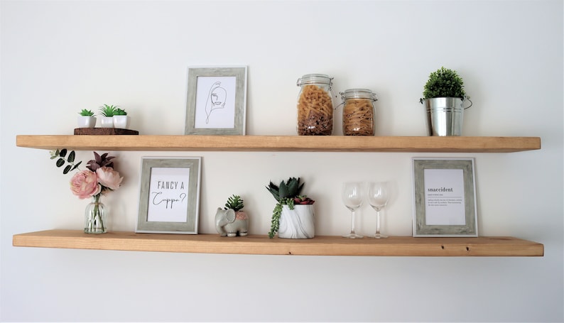 May include: Two wooden shelves with various items on them, including a framed print that says "Fancy a Capp?", a small elephant figurine, two glass jars filled with pasta, two wine glasses, and several small potted plants.