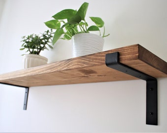 May include: A wooden shelf with black metal brackets. The shelf is stained a dark brown color and has a natural wood grain. Two potted plants are on the shelf.