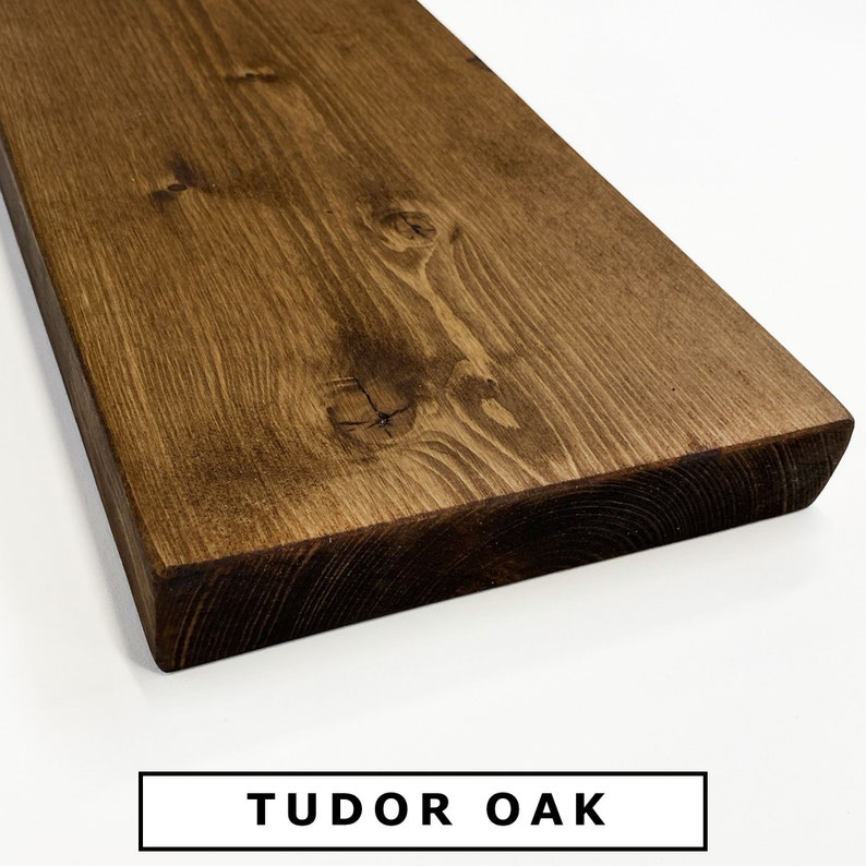 Op de afbeelding: Close-up van een donkerbruin houten plank met een gladde, gebeitst afwerking. De houtnerf is zichtbaar en de plank heeft een licht afgeronde rand. De tekst "TUDOR OAK" is onder de plank gedrukt.