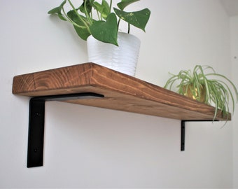 May include: A wooden shelf with black metal brackets. The shelf is stained a dark brown color and has a natural wood grain. Two potted plants are on the shelf.
