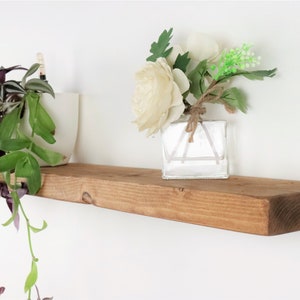 K&ouml;nnte beinhalten: Ein rustikales Holzregal mit einer nat&uuml;rlichen braunen Oberfl&auml;che. Das Regal ist mit einem wei&szlig;en Keramiktopf mit einer h&auml;ngenden Pflanze und einer Glasvase mit wei&szlig;en Blumen dekoriert.