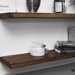 Op de afbeelding: Twee donkerbruine houten planken gemonteerd aan een witte muur. De bovenste plank heeft een klein decoratief object erop. De onderste plank heeft een witte kaars pot en een stapel boeken erop. De tekst "Chill House Hand Crafted Furniture" is zichtbaar in de linkerbovenhoek van de afbeelding. De tekst "WALNUT" is zichtbaar in de rechterbenedenhoek van de afbeelding.