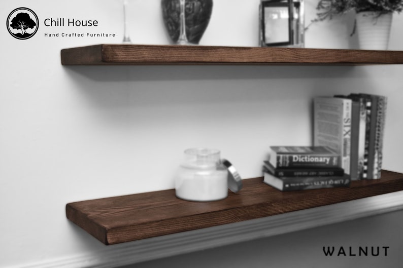 Op de afbeelding: Twee donkerbruine houten planken gemonteerd aan een witte muur. De bovenste plank heeft een klein decoratief object erop. De onderste plank heeft een witte kaars pot en een stapel boeken erop. De tekst "Chill House Hand Crafted Furniture" is zichtbaar in de linkerbovenhoek van de afbeelding. De tekst "WALNUT" is zichtbaar in de rechterbenedenhoek van de afbeelding.