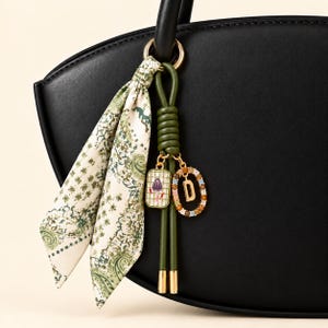 Pode incluir: Uma mala preta com um conjunto de pendentes decorativos presos à pega. O pendente inclui um cordão verde com nós, um pendente retangular com um motivo floral e um pendente circular com a letra D e pedras coloridas.