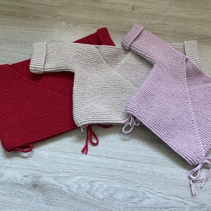 Op de afbeelding: Drie gebreide babyvestjes in rood, beige en lavendel. Elk vestje heeft lange mouwen en een V-hals. De vestjes liggen op een houten oppervlak. Ze hebben striksluitingen aan de onderkant.