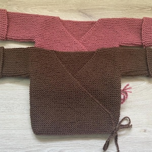Op de afbeelding: Twee gebreide babywikkelvesten. Eén is stoffig roze van kleur, de andere is diepbruin. Beide vesten hebben lange mouwen met omgeslagen manchetten en striksluitingen. Op een licht houten oppervlak.