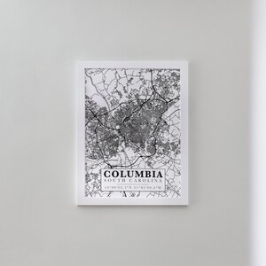 May include: A black and white map print of Columbia, South Carolina, in a white frame. The map shows the city streets and landmarks. The text "COLUMBIA SOUTH CAROLINA 34°00'04.3"N 81°02'00.6"W" is printed below the map.
