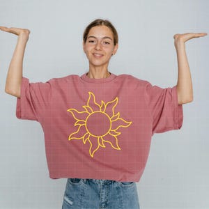 May include: A dusty rose oversized t-shirt featuring a bright yellow graphic of a stylized sun with swirling rays centered on the chest. The garment has short sleeves and a crew neckline, providing a relaxed fit for casual wear.
