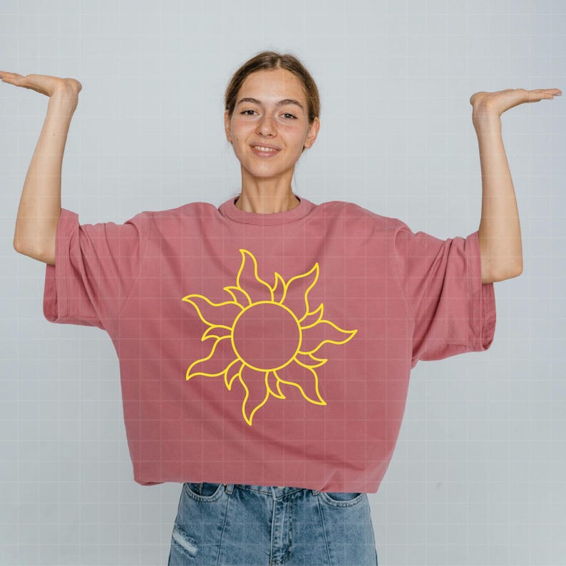 May include: A dusty rose oversized t-shirt featuring a bright yellow graphic of a stylized sun with swirling rays centered on the chest. The garment has short sleeves and a crew neckline, providing a relaxed fit for casual wear.