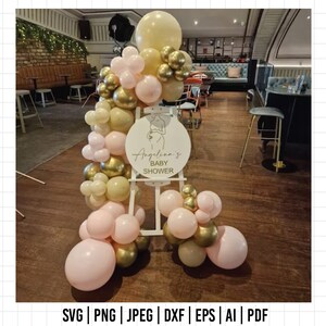 Może przedstawiać: Okrągły biały znak z napisem "Angelion's Baby Shower" na drewnianym stojaku ozdobionym girlandą z r&oacute;żowych, złotych i kremowych balon&oacute;w.