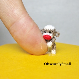 May include: A tiny crocheted monkey with a white head, brown body, and red nose. The monkey is sitting on a yellow surface and is very small, as shown by the finger in the background.