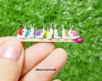 May include: A line of seven miniature crocheted snails in various colors, including red, purple, yellow, green, orange, and pink, are lined up on a matchstick.