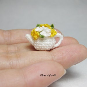 Peut inclure: Une minuscule théière blanche crochetée, ornée d'une couronne de fleurs jaunes et blanches et de feuilles vertes. La théière miniature est dotée d'une anse et d'un bec verseur. Le texte "ObscurelySmall" est visible en bas à droite.