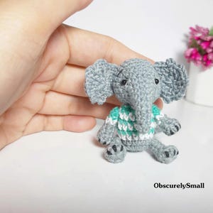 May include: A small, gray crocheted elephant figurine featuring large ears, a trunk, and a white and teal striped sweater. The elephant has black bead eyes and black stitched details on its feet. The text ObscurelySmall is visible.