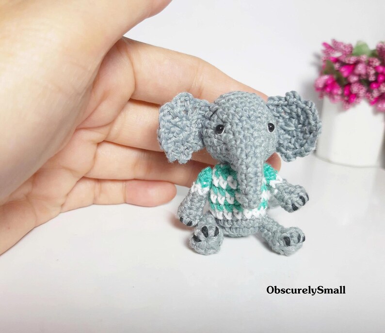 May include: A small, gray crocheted elephant figurine featuring large ears, a trunk, and a white and teal striped sweater. The elephant has black bead eyes and black stitched details on its feet. The text ObscurelySmall is visible.