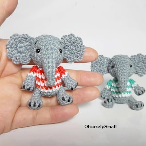 May include: Two small crocheted gray elephant figurines. One elephant wears a red and white striped shirt, and the other wears a teal and white striped shirt. Both have large ears, trunks, and black stitched eyes. The text ObsurelySmall is visible.