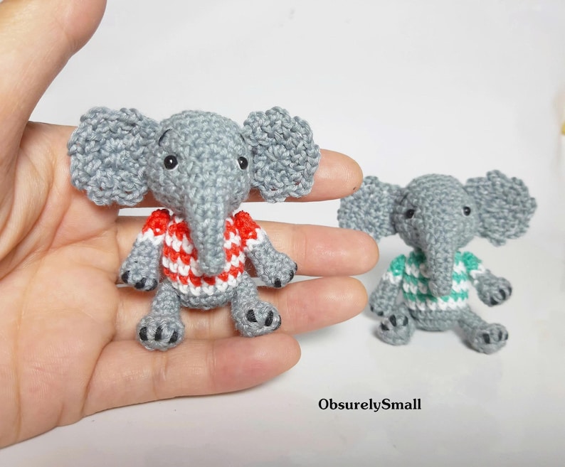May include: Two small crocheted gray elephant figurines. One elephant wears a red and white striped shirt, and the other wears a teal and white striped shirt. Both have large ears, trunks, and black stitched eyes. The text ObsurelySmall is visible.