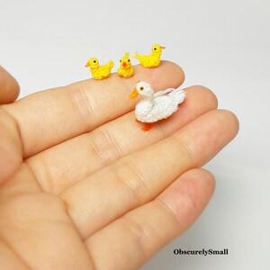 May include: A white crocheted duck with orange feet and a yellow beak is surrounded by three smaller yellow crocheted ducks. The ducks are sitting on a hand.