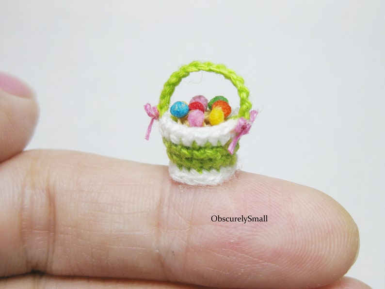 Pu&ograve; includere: Un piccolo cestino all'uncinetto pieno di uova di Pasqua colorate. Il cestino &egrave; verde e bianco con un manico rosa e si trova su un dito.