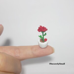 May include: A miniature crocheted red rose in a white pot, held on a fingertip. The flower has green leaves and stem, with a small red heart-shaped accent. The pot is small and round, and the overall effect is delicate and charming.