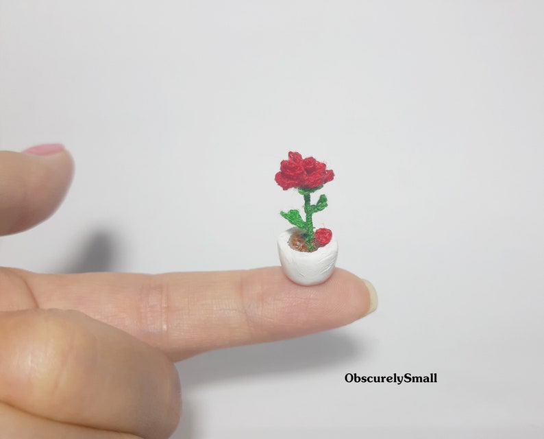 May include: A miniature crocheted red rose in a white pot, held on a fingertip. The flower has green leaves and stem, with a small red heart-shaped accent. The pot is small and round, and the overall effect is delicate and charming.