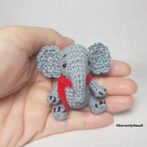 May include: A small gray crocheted elephant figurine featuring large ears, black bead eyes, and a bright red scarf wrapped around its neck. The elephant has black stitched details on its feet and the text ObscurelySmall is visible in the corner.