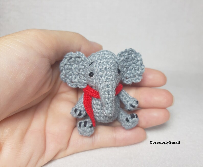 May include: A small gray crocheted elephant figurine featuring large ears, black bead eyes, and a bright red scarf wrapped around its neck. The elephant has black stitched details on its feet and the text ObscurelySmall is visible in the corner.
