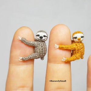 May include: Two miniature crocheted sloths, one grey and one orange, are hanging on fingers. The sloths are very small, about the size of a fingernail. The text "ObscurelySmall" is visible in the bottom centre of the image.