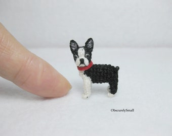 May include: A small crocheted Boston Terrier dog with black and white fur wearing a red collar. The dog is standing on a white surface.