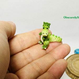May include: A small green crocheted dinosaur toy with white accents. The dinosaur is sitting in the palm of a hand.
