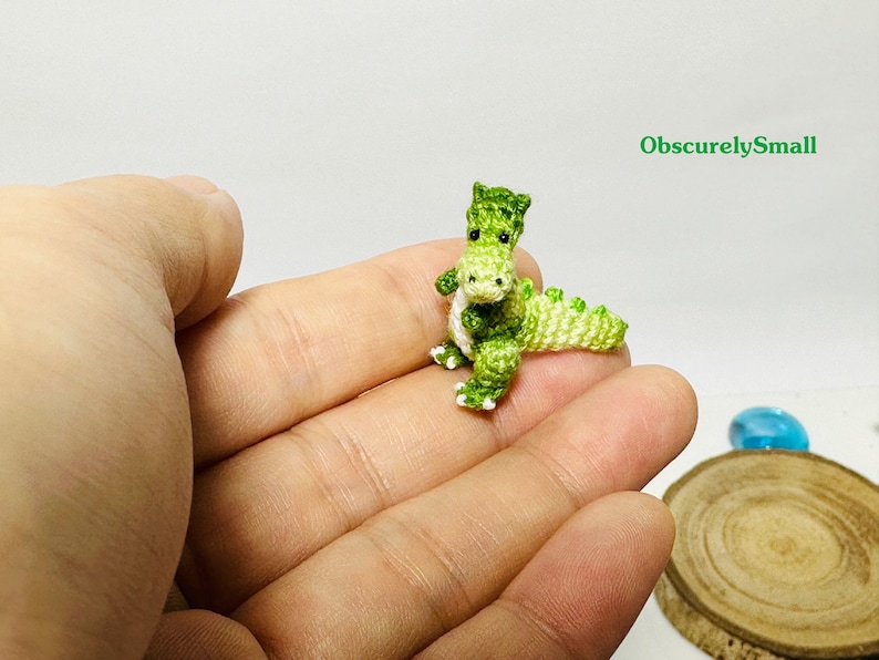 May include: A small green crocheted dinosaur toy with white accents. The dinosaur is sitting in the palm of a hand.