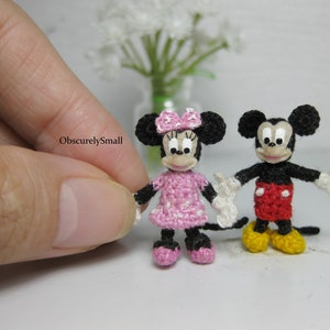 Puede incluir: Un par de figuras de Mickey Mouse y Minnie Mouse de ganchillo. Mickey lleva pantalones cortos rojos y zapatos negros. Minnie lleva un vestido de lunares rosa y zapatos negros.