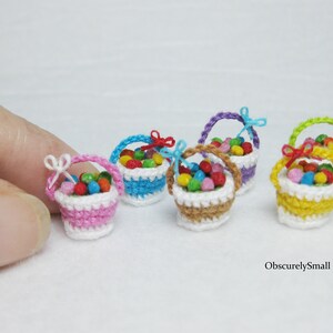 Pu&ograve; includere: Cinque cestini in miniatura all'uncinetto pieni di caramelle colorate. I cestini sono rosa, blu, marrone, viola e giallo. Ogni cestino ha un bordo in crochet bianco e un piccolo fiocco.