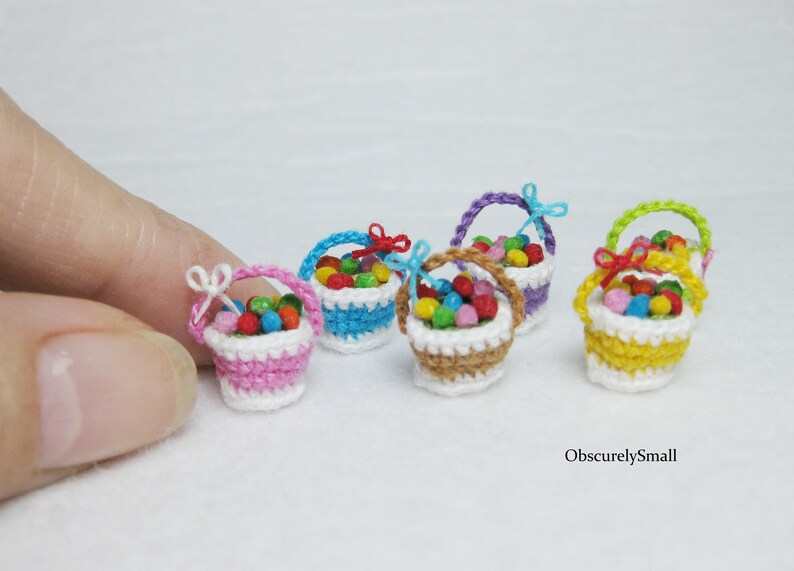 Pu&ograve; includere: Cinque cestini in miniatura all'uncinetto pieni di caramelle colorate. I cestini sono rosa, blu, marrone, viola e giallo. Ogni cestino ha un bordo in crochet bianco e un piccolo fiocco.
