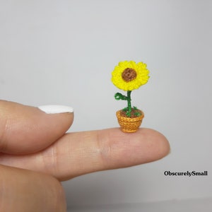 May include: A miniature crocheted sunflower in a small woven pot. The sunflower has bright yellow petals and a brown center. The pot is a warm orange color, and the stem and leaves are green. The text "ObscurelySmall" is in the bottom right corner.