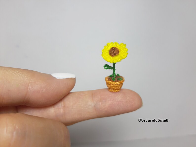 May include: A miniature crocheted sunflower in a small woven pot. The sunflower has bright yellow petals and a brown center. The pot is a warm orange color, and the stem and leaves are green. The text "ObscurelySmall" is in the bottom right corner.