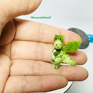 May include: A small green crocheted dragon with white accents and a green crocheted wing. The dragon is sitting on a person's hand.