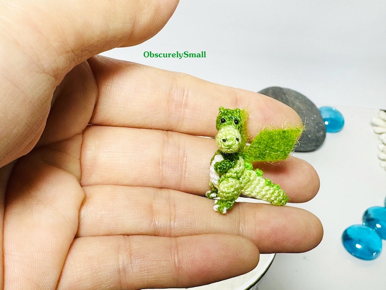 May include: A small green crocheted dragon with white accents and a green crocheted wing. The dragon is sitting on a person's hand.