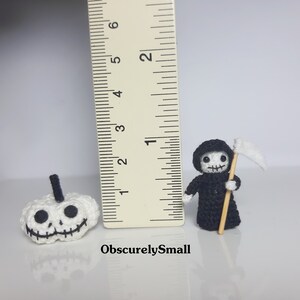 May include: Two crocheted Halloween decorations, a white pumpkin with a black face and a black Grim Reaper with a white scythe. The decorations are about 2 inches tall and are next to a ruler for scale.