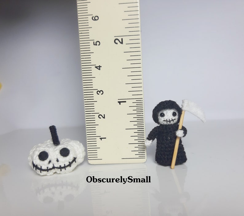 May include: Two crocheted Halloween decorations, a white pumpkin with a black face and a black Grim Reaper with a white scythe. The decorations are about 2 inches tall and are next to a ruler for scale.