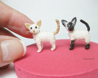 May include: Two crocheted miniature Siamese cats, one with a tan tail, standing on a pink surface. The text "ObscurelySmall" is visible in the bottom right corner.