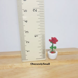 May include: A miniature crocheted red rose in a white ceramic pot. The green stem and leaves support the flower. The pot is approximately 1 inch tall. The text "ObscurelySmall" is visible.